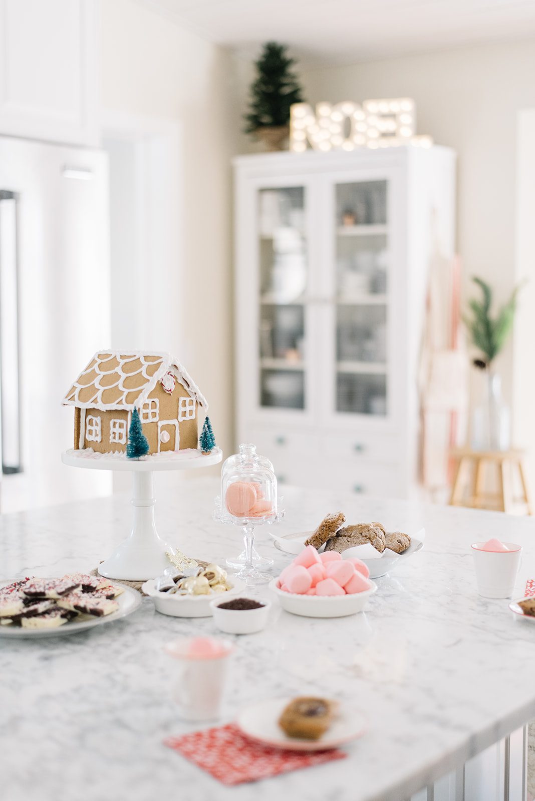gingerbread house sits on a white cake pedestal beside cookies and marshmallows