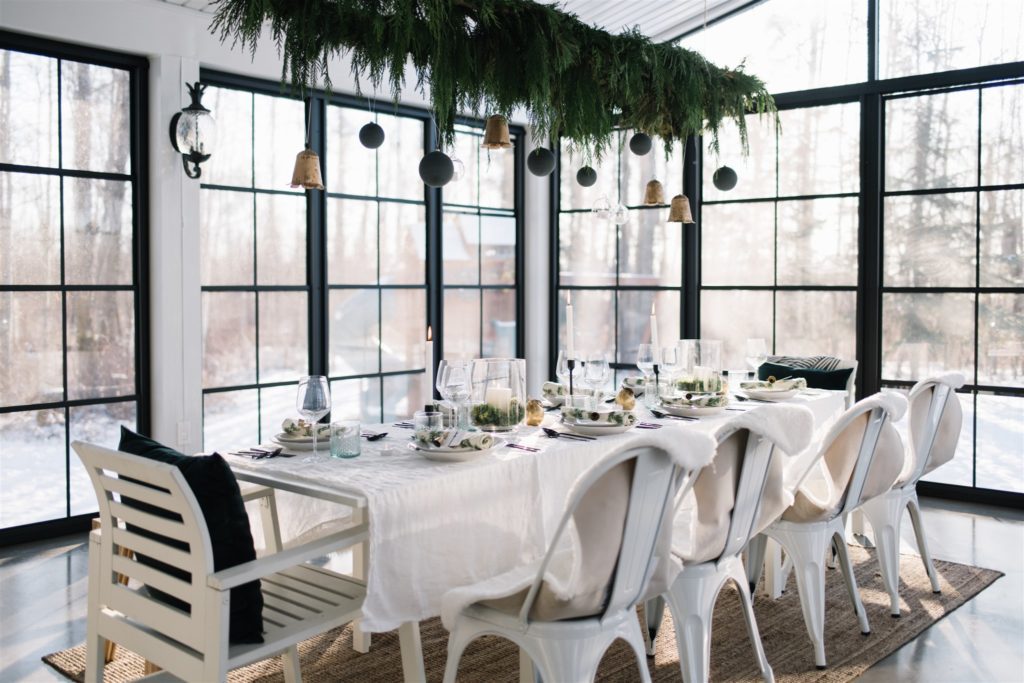 A sun room with a dining table set up for Christmas with greens and Christmas ornaments hanging above