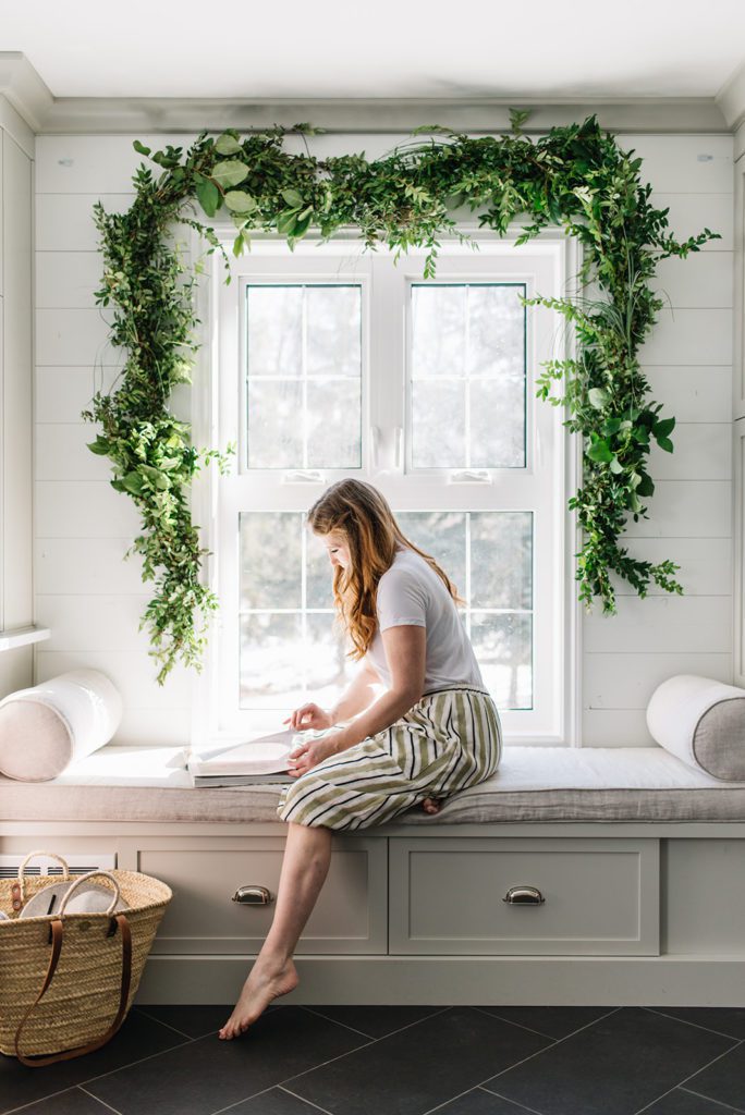 woman sits on bench reading book beneath fresh green garland over window