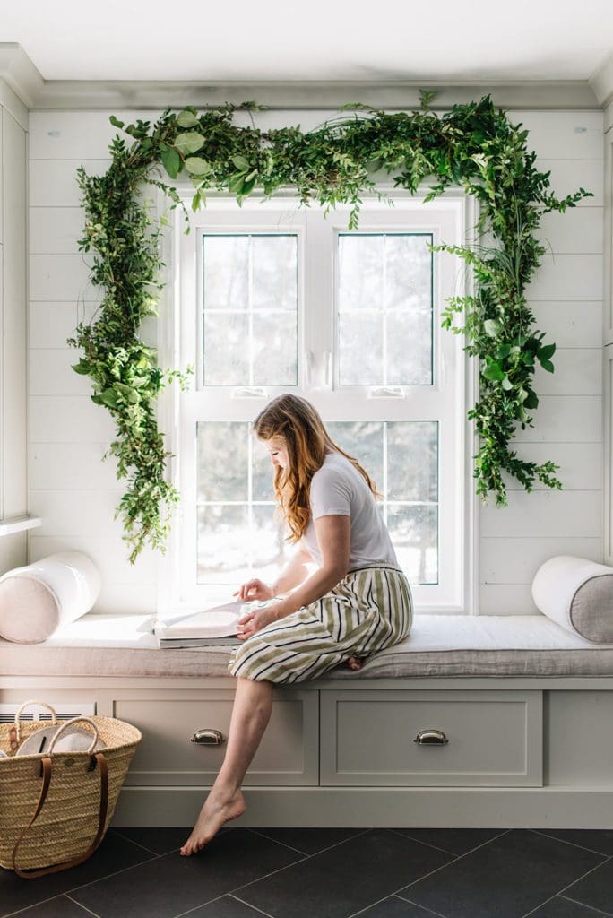 woman sits on bench reading book beneath fresh green garland over window