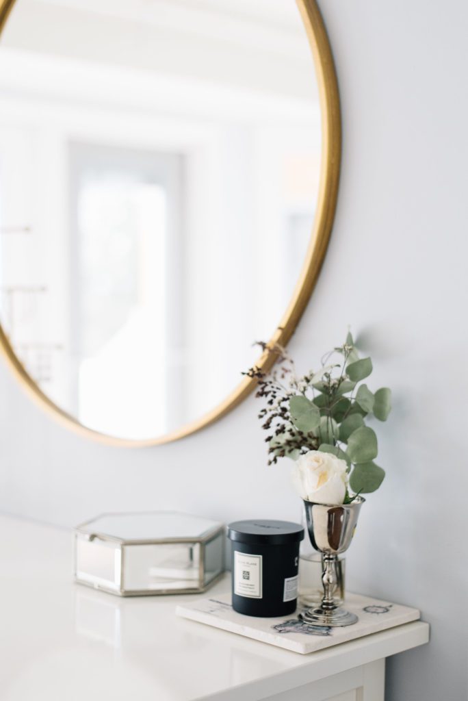 A simple silver vase of fresh flowers sits under a gold framed mirror on a white dresser in a master bedroom