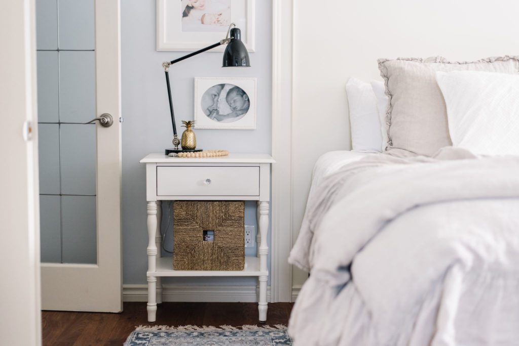 A white bedside table with a basket for storage beside a bed in a small master bedroom