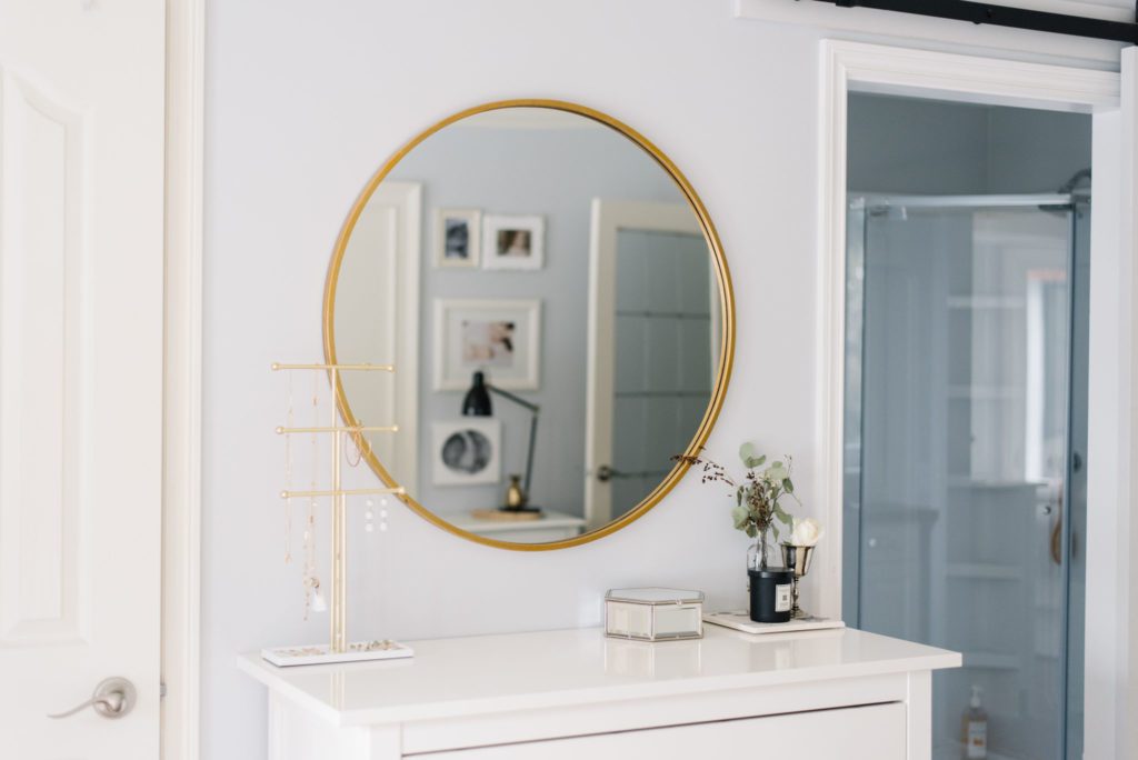 A gold framed round mirror above a white dresser bounces light into a small master bedroom