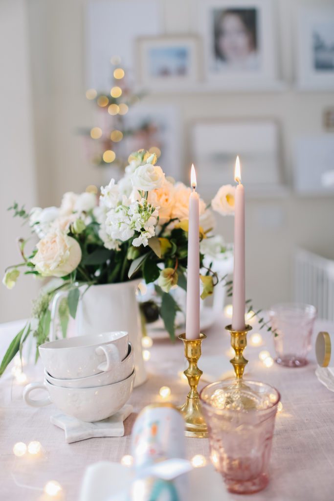 A vase of flowers on a table behind pink candles in brass holders and a stack of white mugs