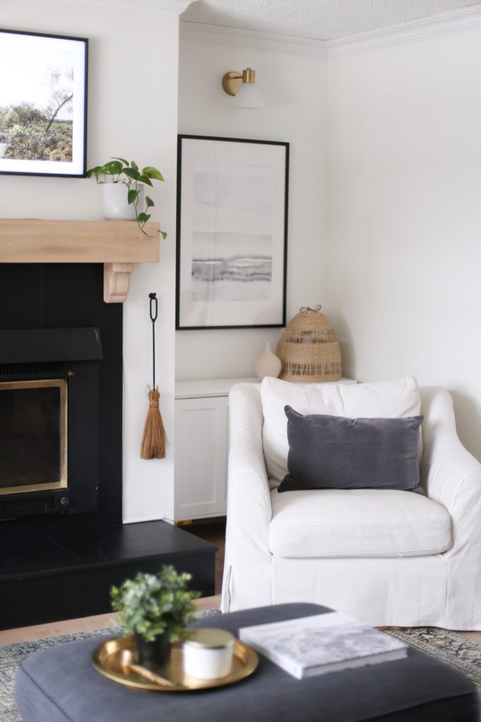 A white armchair nestled beside the wood burning fireplace with white oak mantle in the living room of a 1950s character home