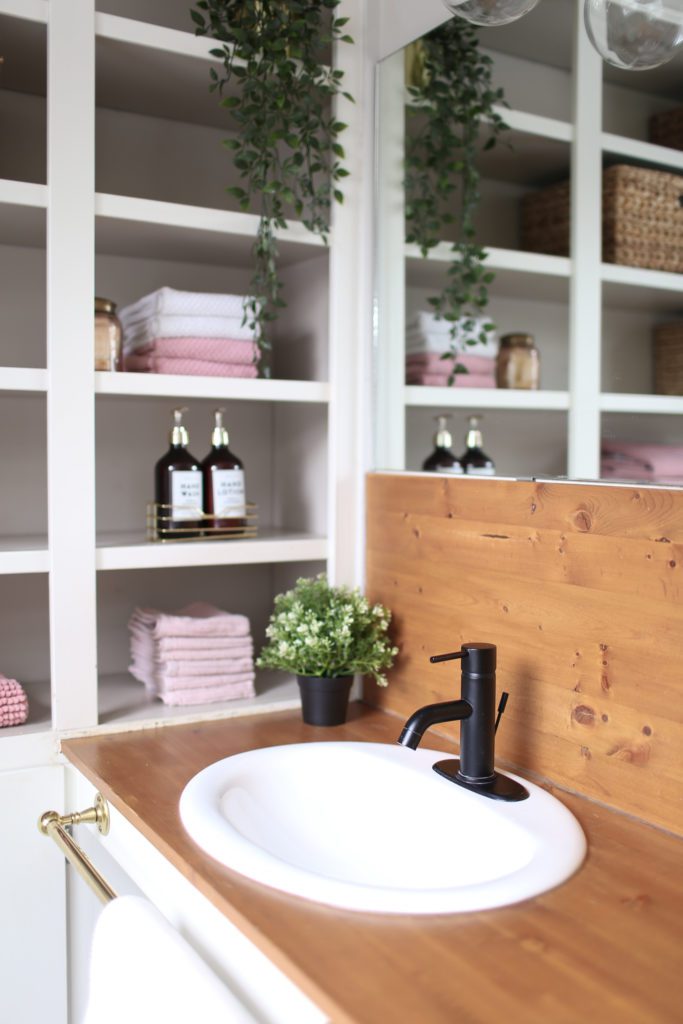 A wooden topped bathroom vanity with open shelves beside
