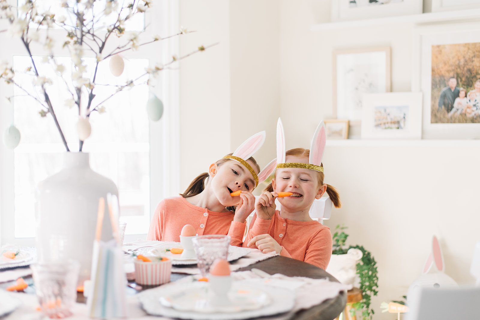 2 little girls with bunny ears sitting at a table set up Easter pretending to eat carrots