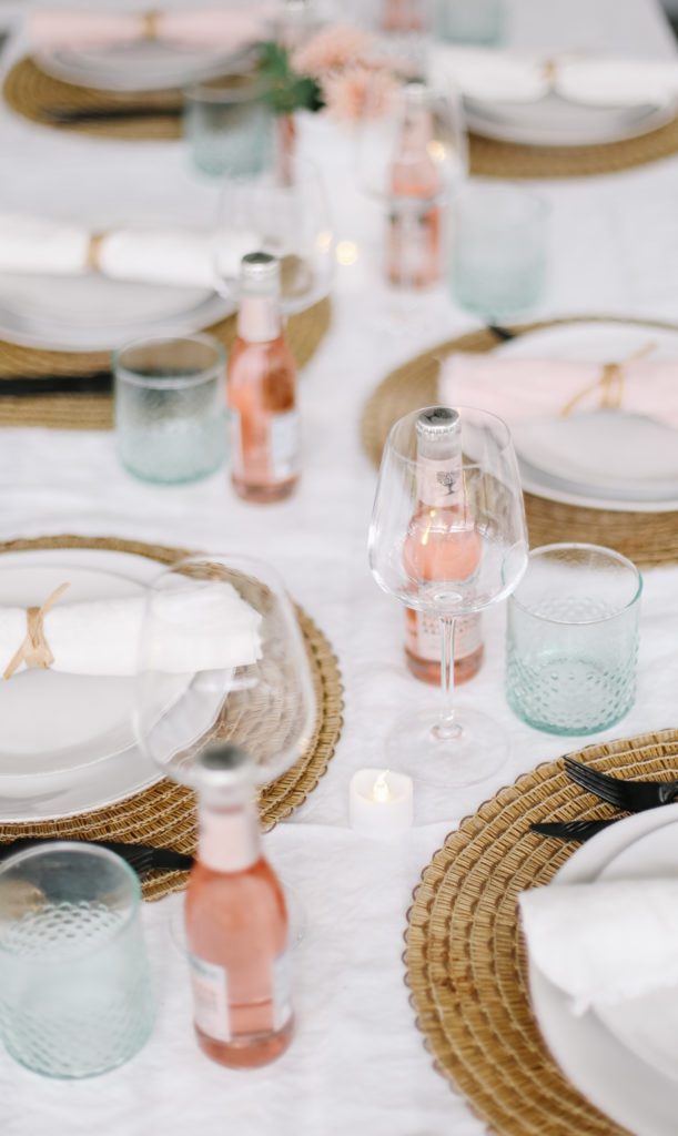 A dining table set with clear wine glasses, green water glasses, pink bottles and seagrass place mats