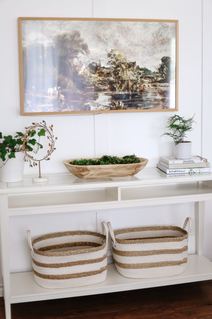 A white console table with plants, books and baskets and a Samsung Frame TV displayed above